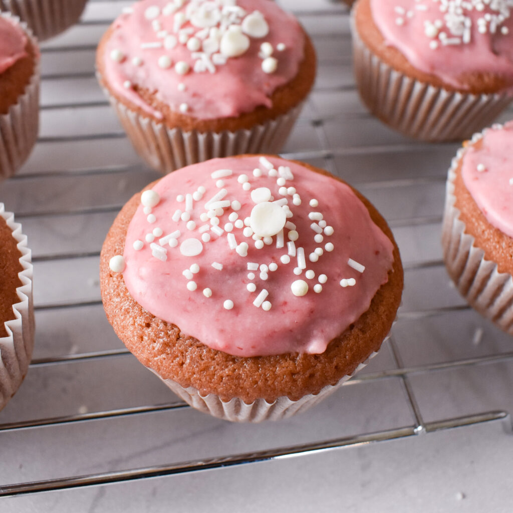 Strawberry cupcakes on cooling rack. Cupcakes are pink with strawberry glaze and white sprinkles. 