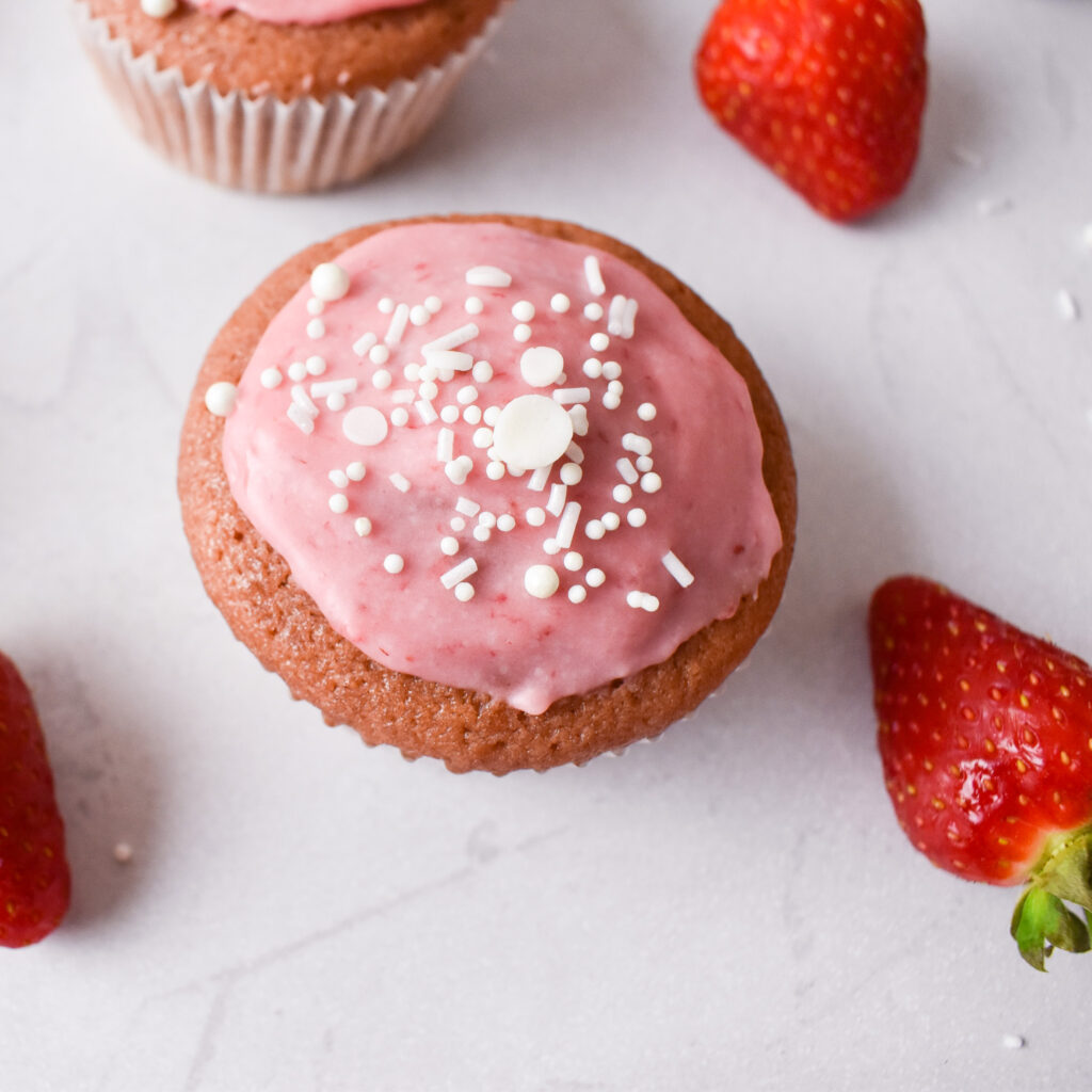 Top view of a strawberry cupcake with pink glaze and white sprinkles.