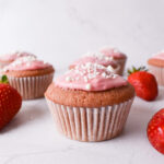 Strawberry cupcake with a pink glaze and white sprinkles. More cupcakes in the background.