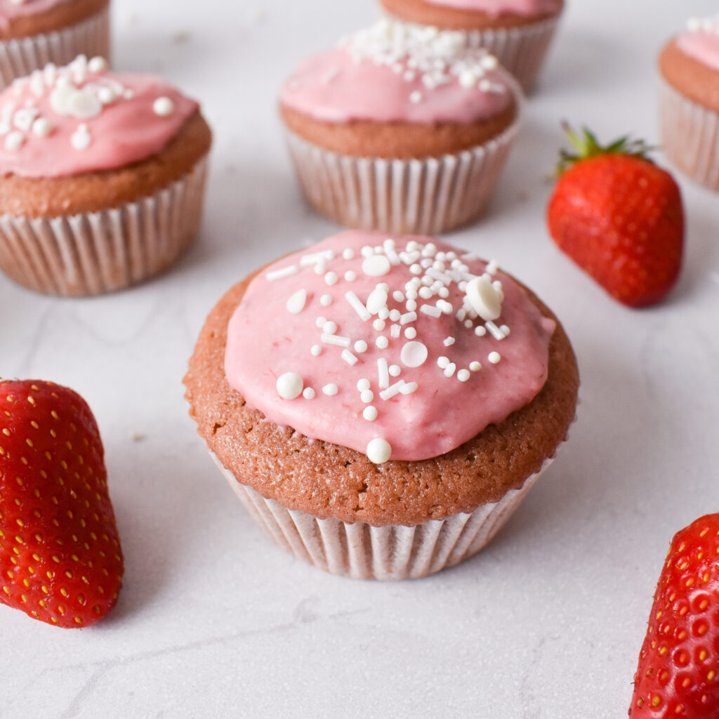 Pink strawberry cupcakes a few inches apart. Cupcakes are topped with pink glazed icing and white sprinkles. 